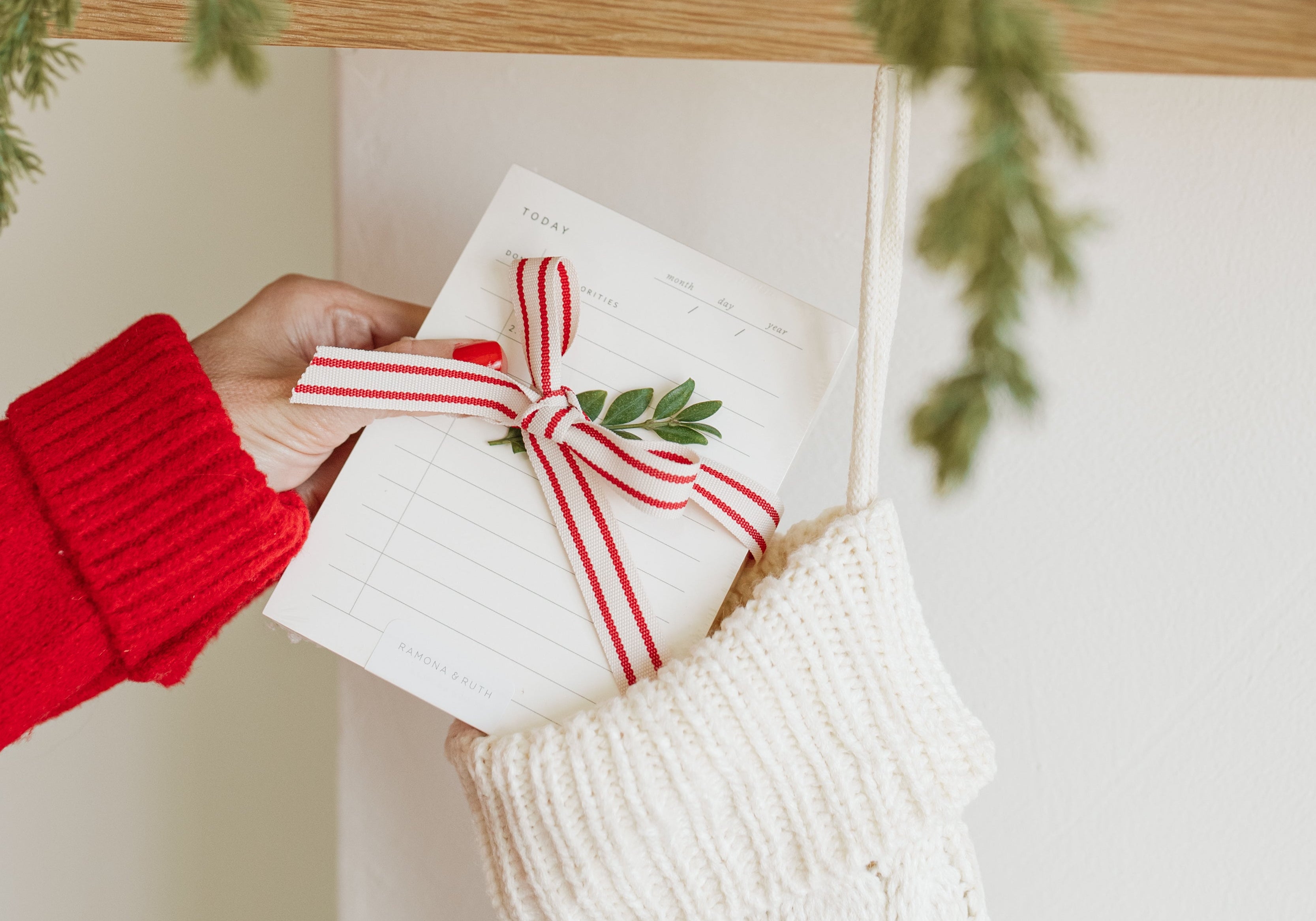 Person holding a notepad gift with a red ribbon in front of a decorated fireplace mantel.