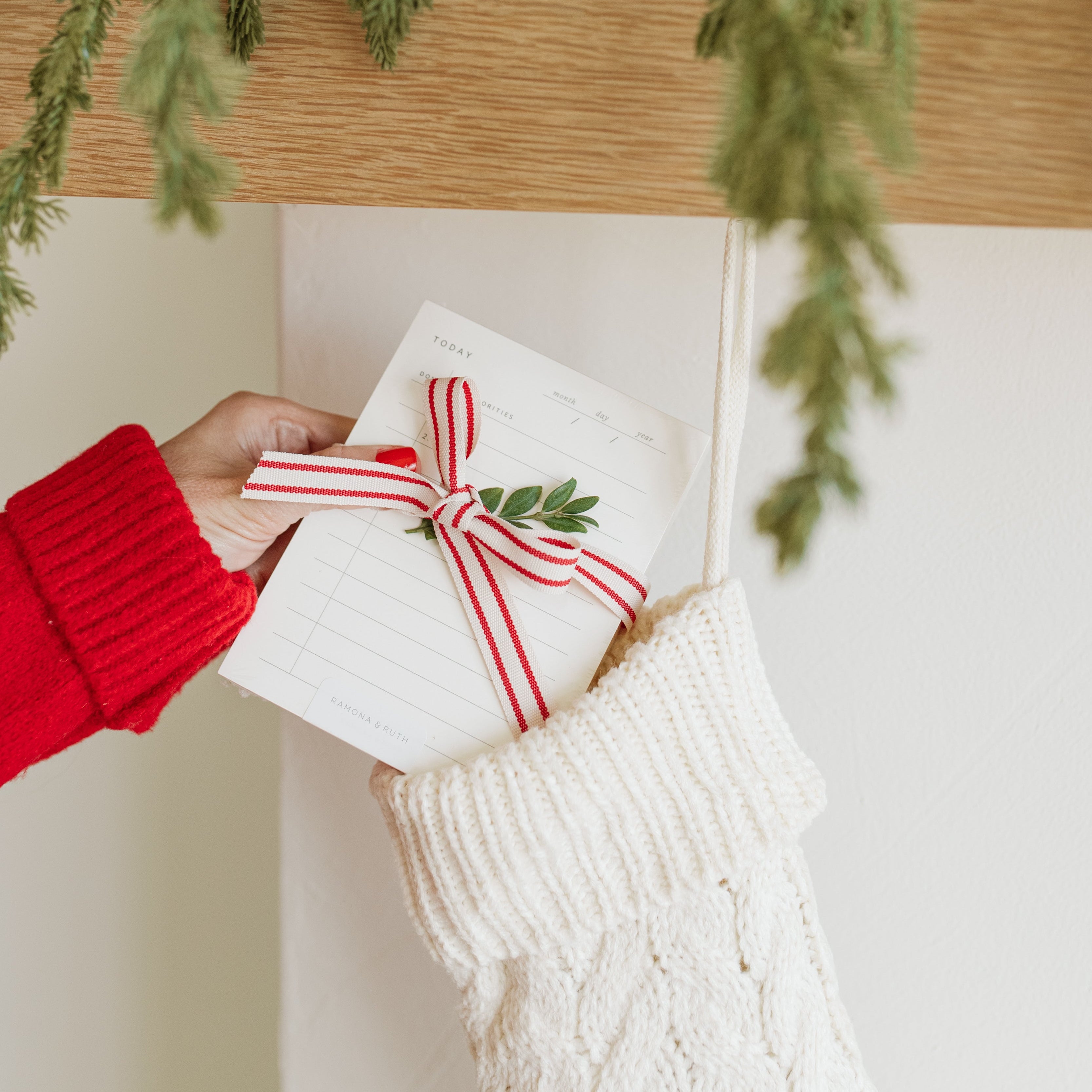 Person holding a notepad gift with a red ribbon in front of a decorated fireplace mantel.